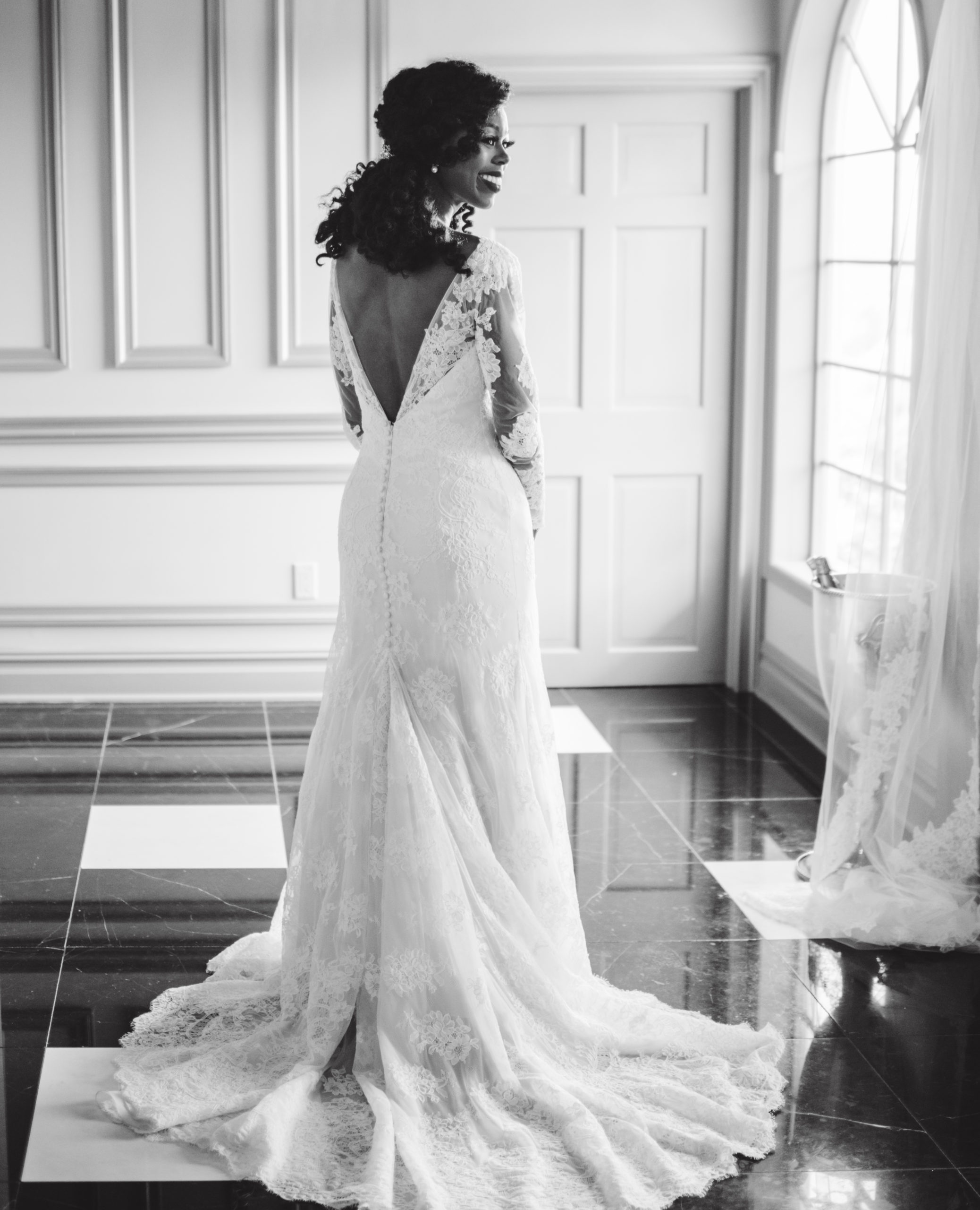 Black and white photo of a beautiful bride standing in her v-back wedding dress next to a window