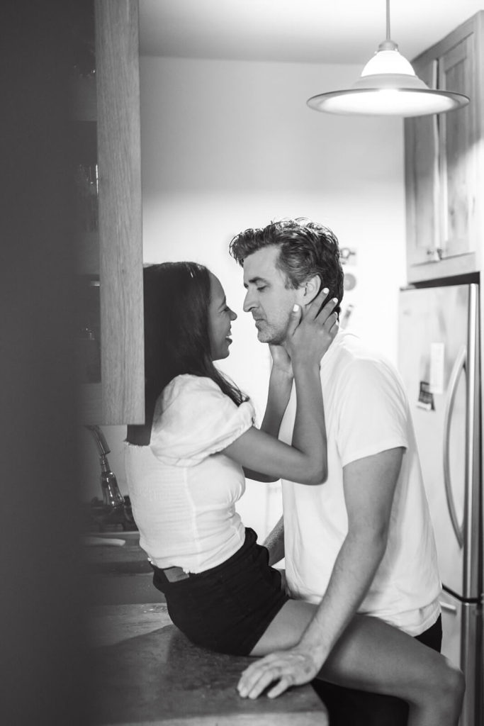brunette woman in white top sits on kitchen countertop with her partner's face in her hands as he leans in for a kiss