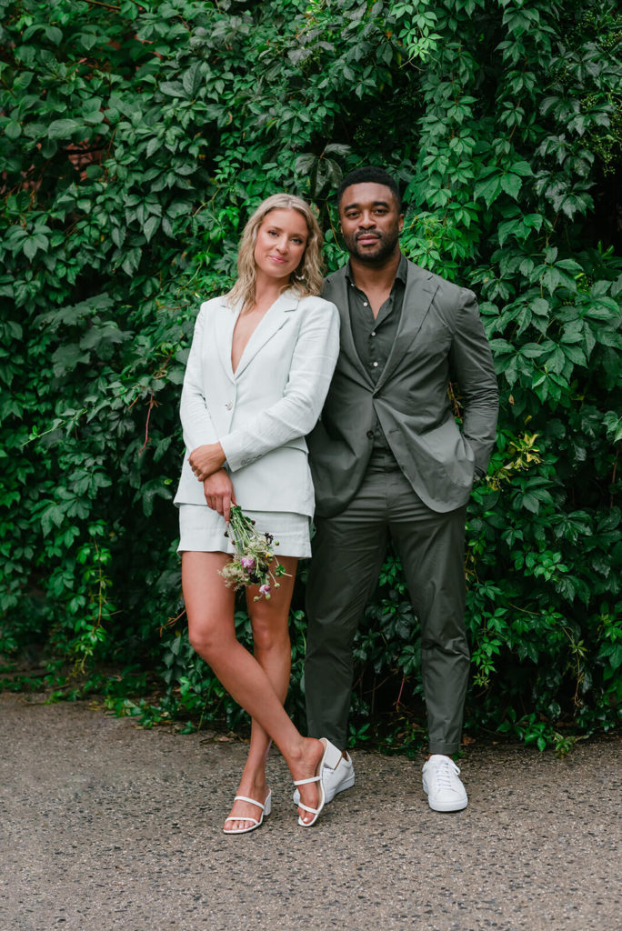 Blonde woman in a white blazer smiles into the camera standing beside her handsome black husband-to-be during their Brooklyn summer engagement session
