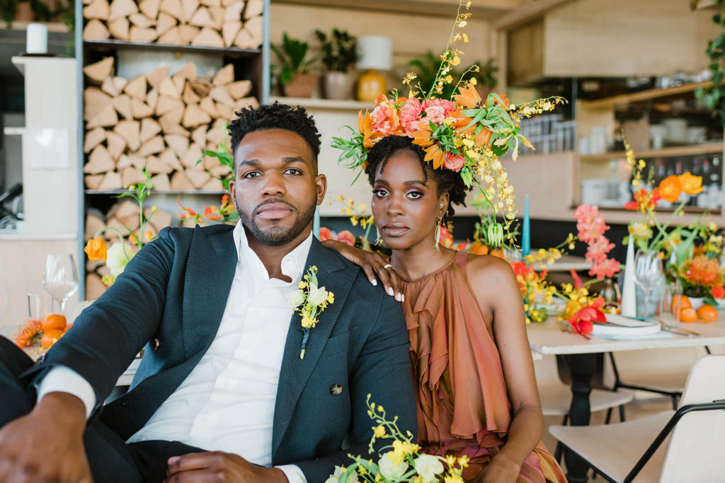black couple leans on one another inside a brightly lit restaurant as the woman wears a stunning flower crown