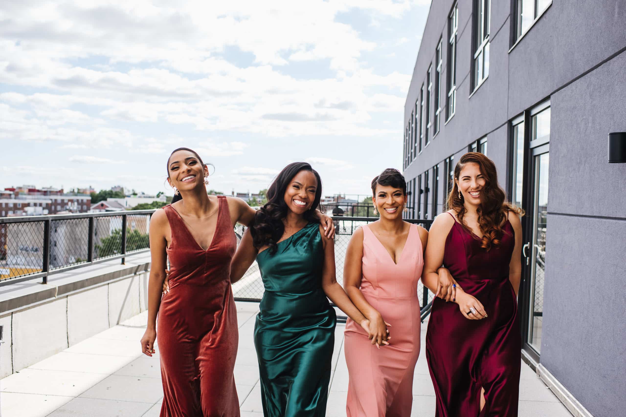 four bridesmaids standing on a deck in jewel-toned dresses as they smile and walk toward the camera