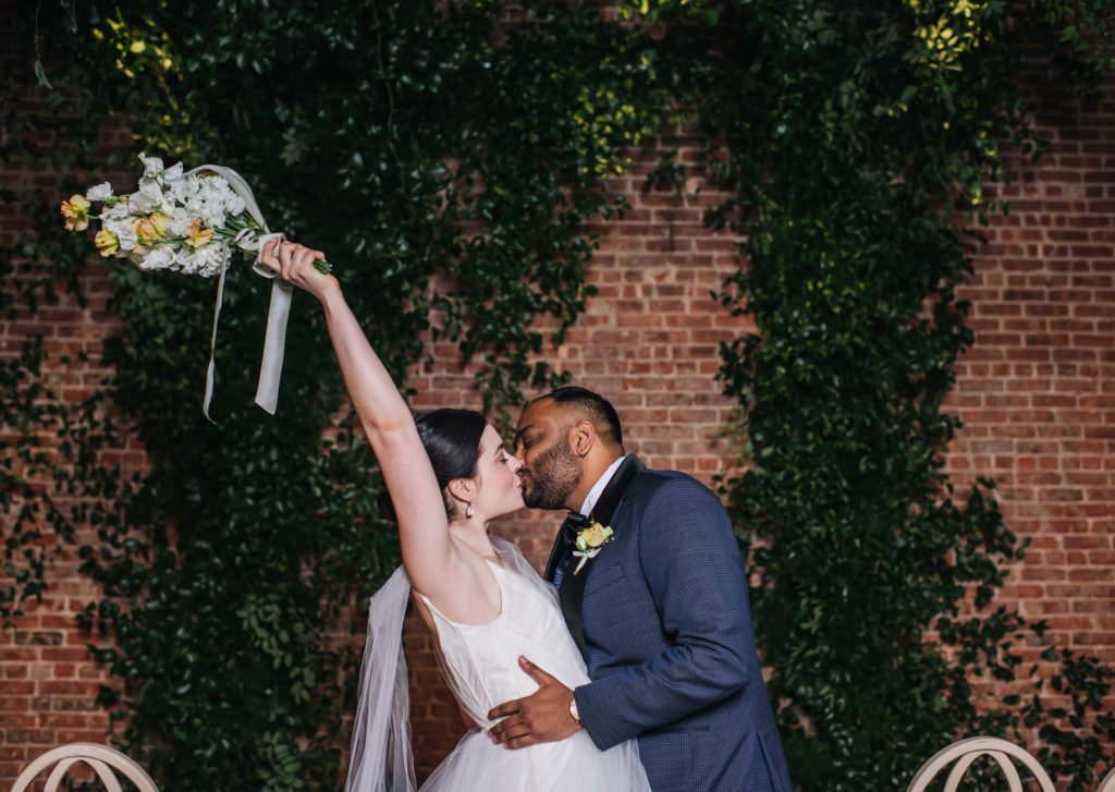 Newlyweds kiss in front of a brick wall outside as the bride holds her bouquet in the air