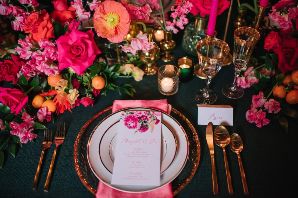 Beautiful stationary rests on top of a stunning pink and orange wedding tablescape full of flowers, candles, and gold accents